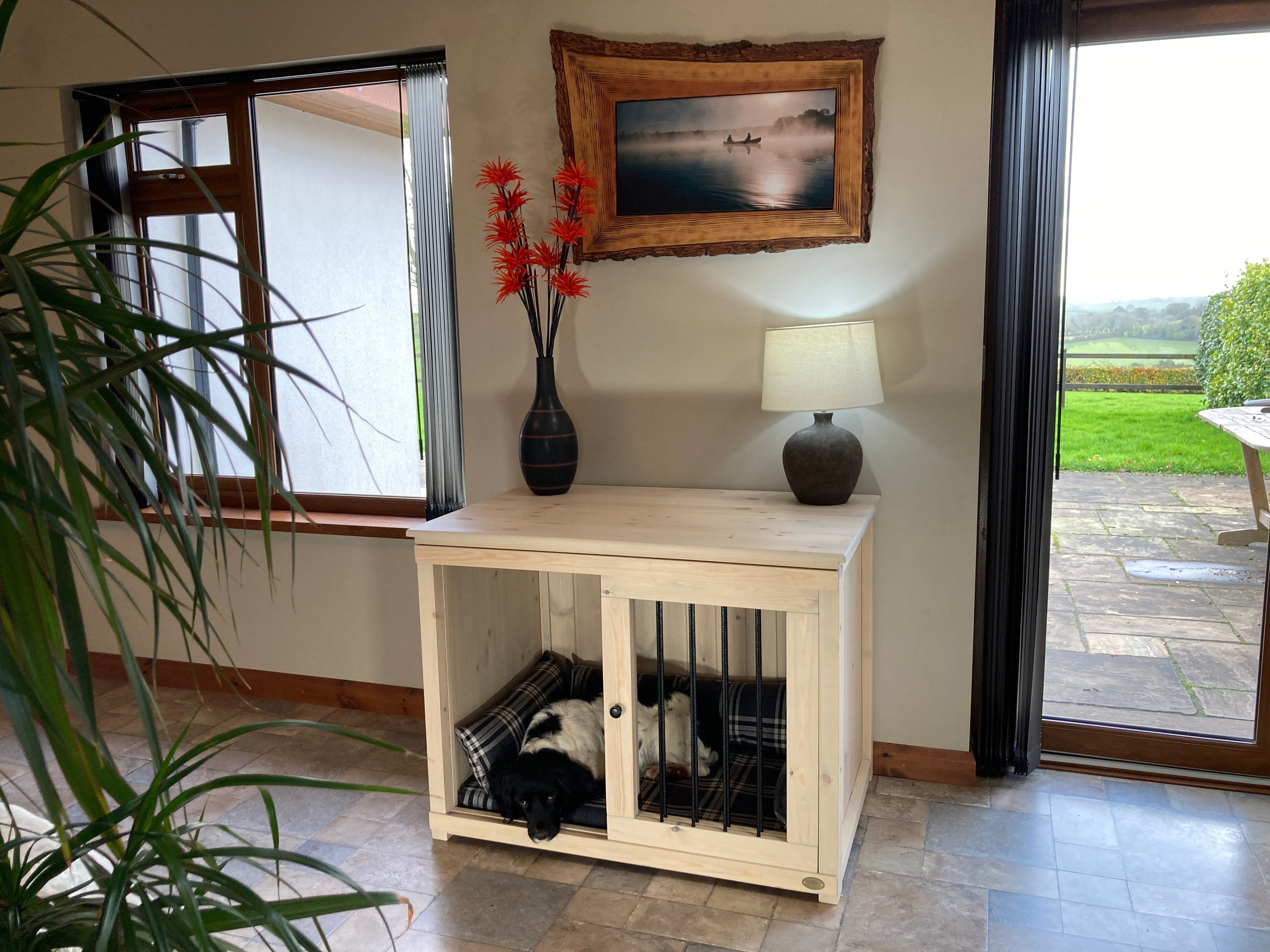 Handmade single wooden dog crate cabinet with black and white dog resting inside in a bright hallway overlooking the countryside